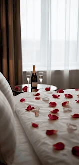 A romantic hotel bed with scattered red rose petals, a bottle of champagne, and two fluted glasses by a sunlit window, waiting for a special moment.