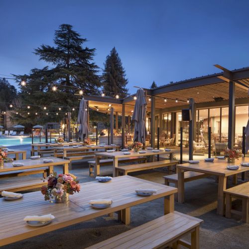 Outdoor dining area with wooden tables, benches, string lights, and floral arrangements. A pool and trees are visible in the background.