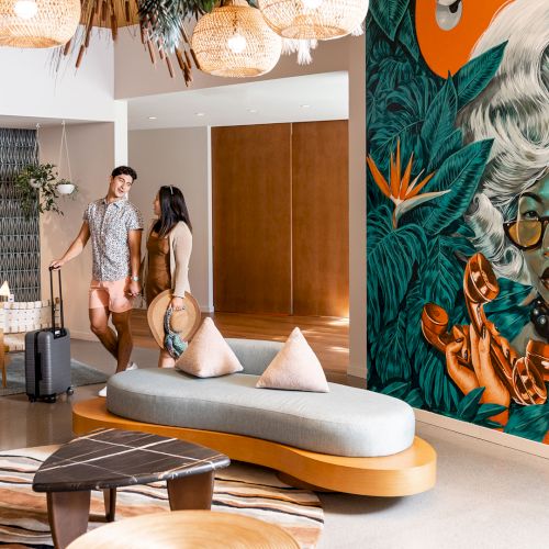 A stylish hotel lobby with modern decor, a vibrant mural, and two people with luggage walking near the seating area.