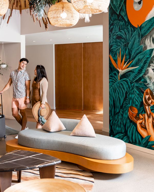 A stylish hotel lobby with modern decor, a vibrant mural, and two people with luggage walking near the seating area.