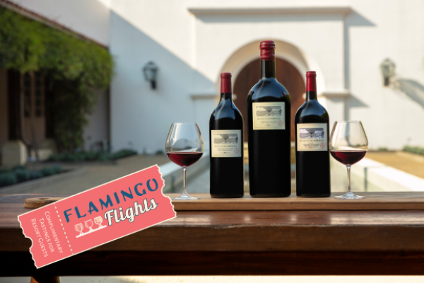 Three wine bottles and two glasses on a wooden table, with a ticket labeled "Flamingo Flights" in an outdoor setting.