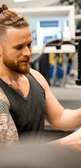 A person with a tattooed arm is adjusting gym equipment in a fitness center, wearing a black tank top.