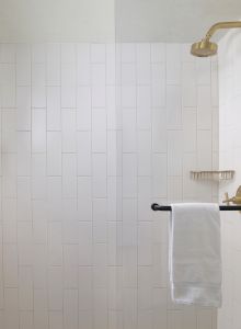 A clean, tiled shower features a gold showerhead, wall-mounted dispensers, a towel rack with a towel, and a wire shelf.