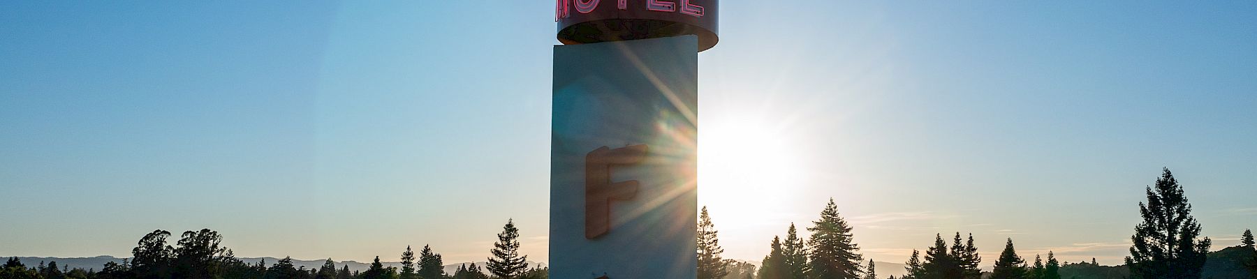 A tall sign with a flamingo and "HOTEL" on top, "FLA" below, set against a sunrise or sunset sky with trees and rooftops in the background.