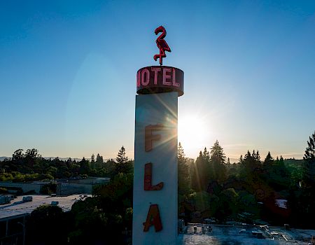A tall sign with a flamingo and "HOTEL" on top, "FLA" below, set against a sunrise or sunset sky with trees and rooftops in the background.