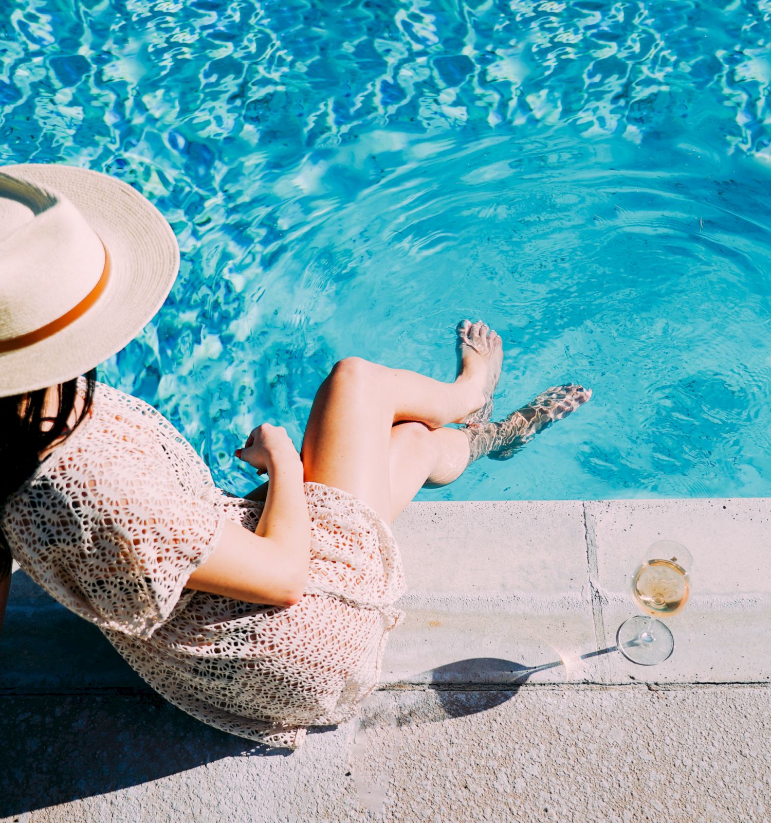 A person in a sun hat sits by a pool with feet in the water, holding a drink, next to a glass of white wine on the edge.