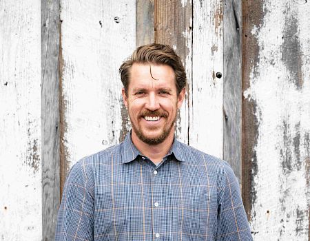 A person stands smiling in front of a rustic wooden wall, wearing a checkered shirt and jeans.