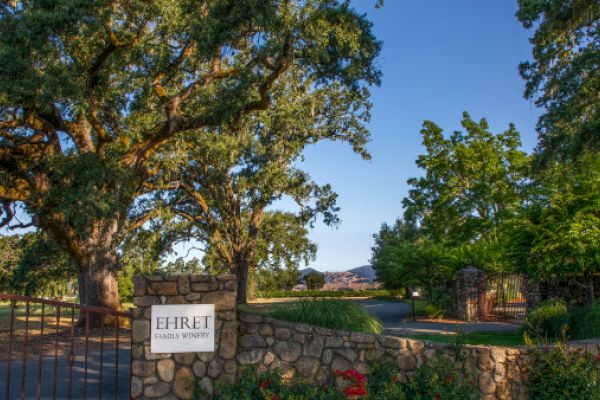 The image shows a scenic winery entrance with large trees and a stone gate, with a sign reading "Ehret Family Winery."