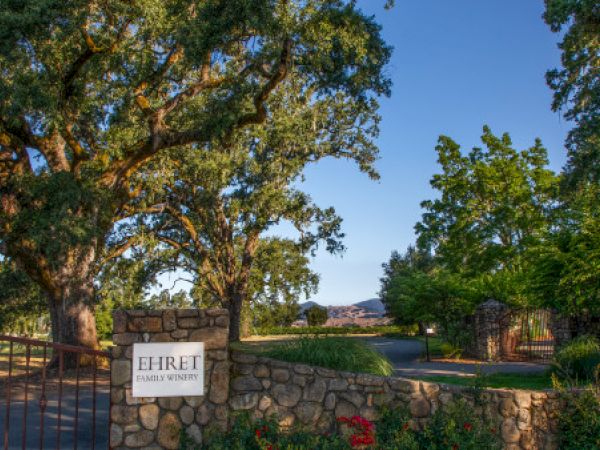 The image shows a scenic winery entrance with large trees and a stone gate, with a sign reading "Ehret Family Winery."