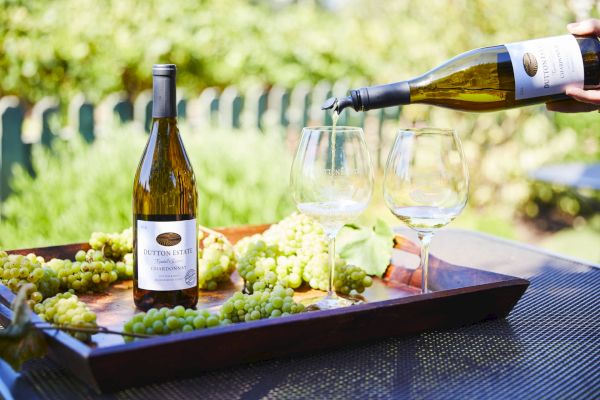 A tray with wine bottles, glasses, and grapes is set outdoors, with wine being poured into a glass.