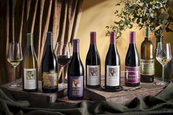 The image shows a variety of wine bottles with different labels, two filled wine glasses, and some greenery and drapery around them.