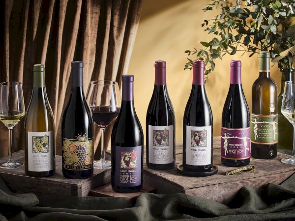 The image shows a variety of wine bottles with different labels, two filled wine glasses, and some greenery and drapery around them.