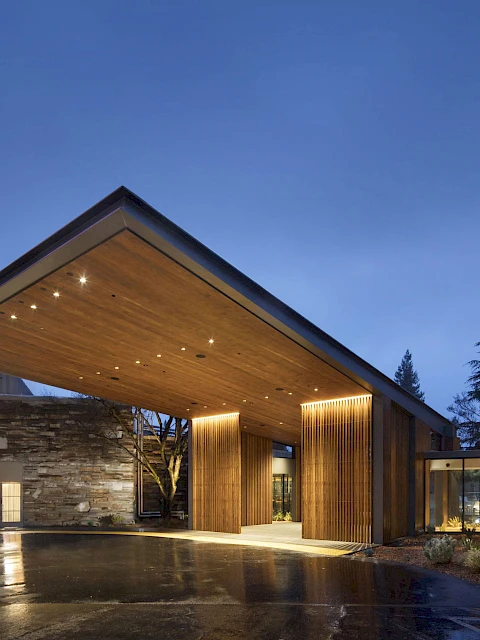 A modern building with a large, illuminated wooden canopy and glass walls, set against a twilight sky with surrounding trees and landscaping.