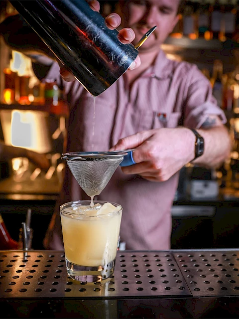 A bartender is pouring a yellow cocktail through a strainer into a glass on a bar counter.