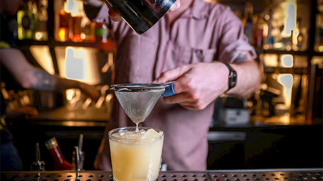 A bartender is pouring a yellow cocktail through a strainer into a glass on a bar counter.