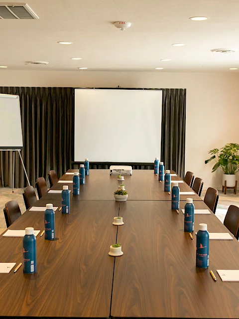 A conference room with a long table, chairs, notepads, water bottles, a whiteboard, and a projector screen is shown.