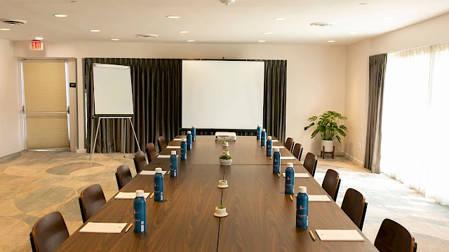 A conference room with a long table, chairs, notepads, water bottles, a whiteboard, and a projector screen is shown.