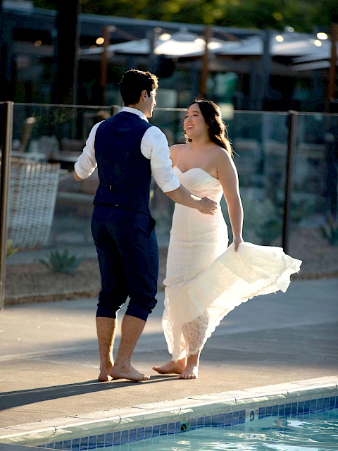A couple is joyfully dancing near a pool, with the woman wearing a white dress and the man in a vest and trousers.