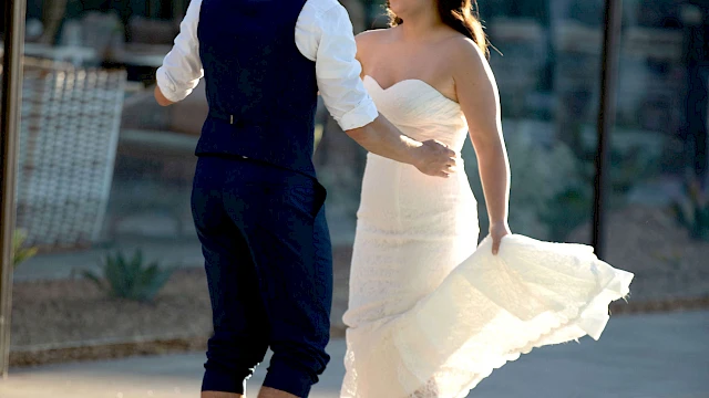 A couple is joyfully dancing near a pool, with the woman wearing a white dress and the man in a vest and trousers.