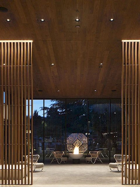 A modern architectural space with wooden vertical slats, ceiling lights, and hanging chairs leads to an outdoor area at twilight.