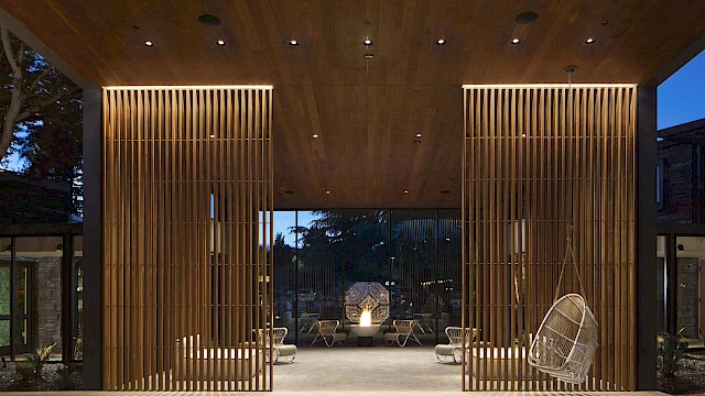 A modern architectural space with wooden vertical slats, ceiling lights, and hanging chairs leads to an outdoor area at twilight.