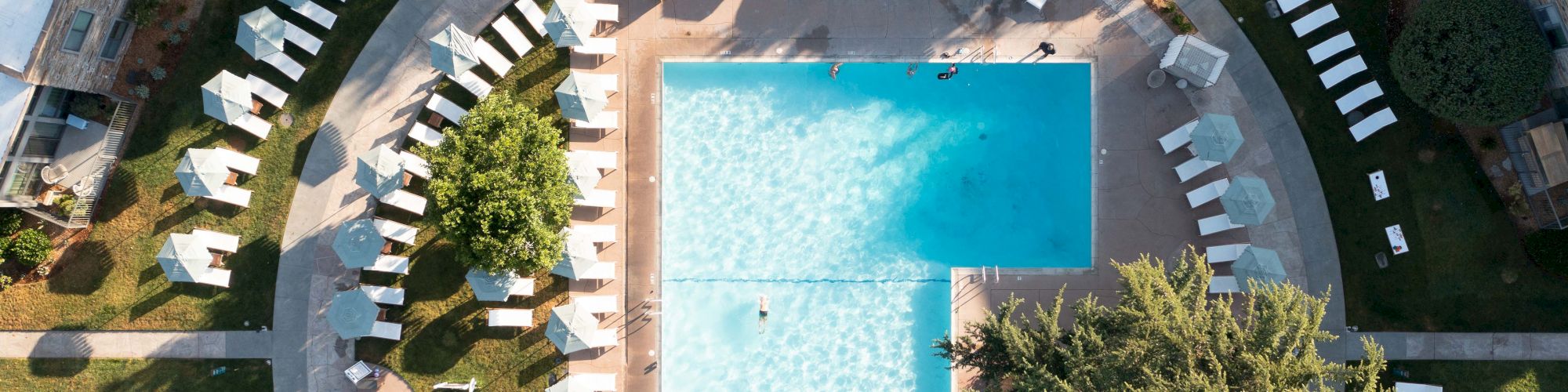 Aerial view of a sunny resort pool with surrounding patios, umbrellas, lounge chairs, and a curved walkway ringing the area.