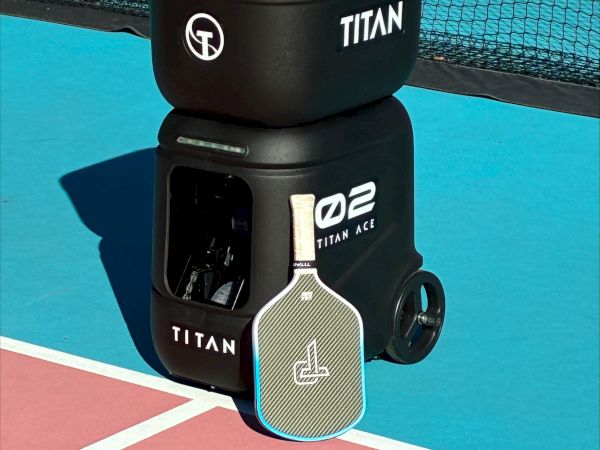 A black Titan tennis ball hopper is stacked on a hopper ball machine, full of yellow balls, on a blue and pink court with a net in the background.