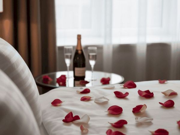 A romantic hotel bed with rose petals scattered across white sheets, a bottle of champagne and two glasses near a window.