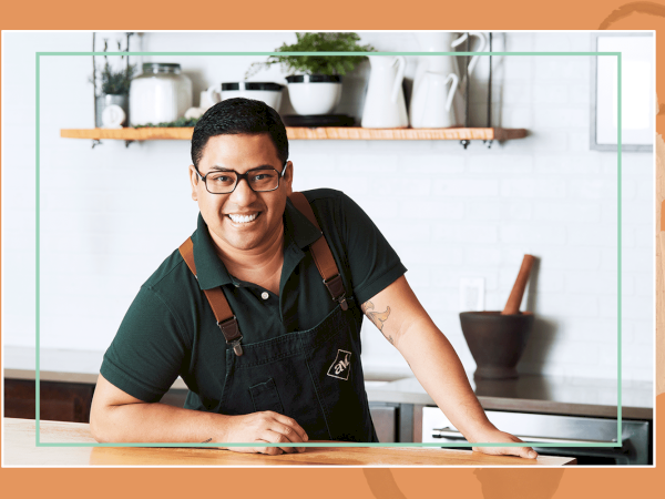 A smiling handyman or barista leaning on a counter in a kitchen, wearing a dark polo and apron.
