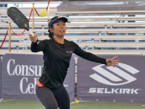 A female tennis player serves during a match on an outdoor court, mid-swing with a racquet, wearing black athletic gear and cap, seated banners in the background.