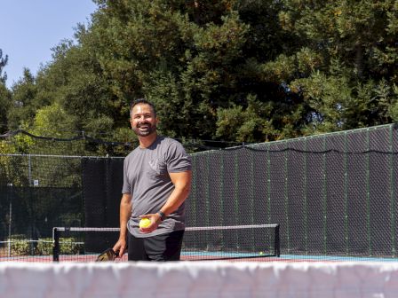 A man stands on a tennis court near the net, holding a tennis ball, with trees and a fence in the background.