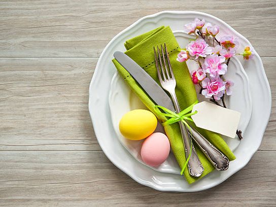 A white plate holds a green napkin, silverware, two pastel eggs, a small block of cheese, pink flowers, and a tiny pink flower on a light wooden table.