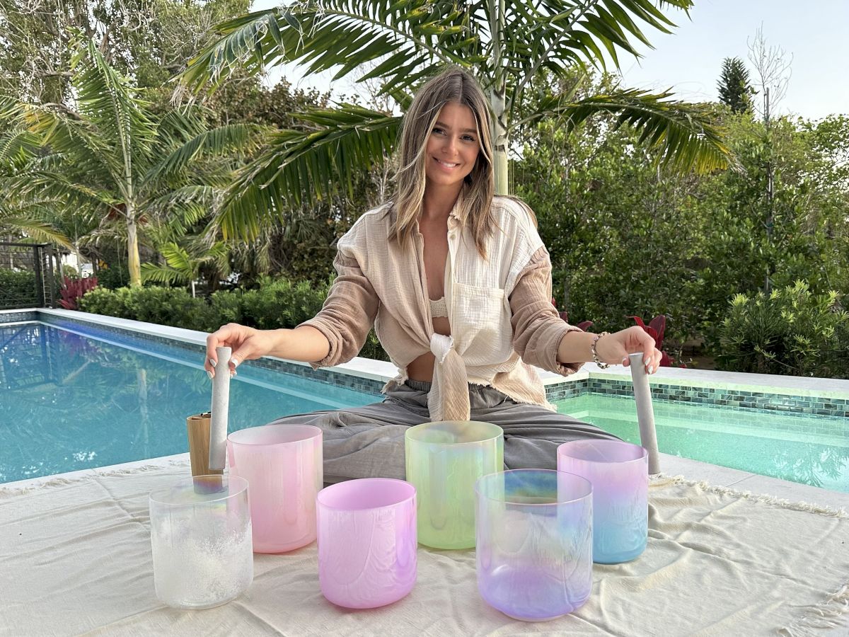 A woman by a pool with colorful glass candle holders in front, adjusting two wooden sticks, sunlight and palm trees in the background.