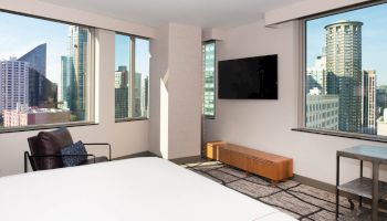 The image depicts a modern hotel room with large windows offering a cityscape view, a wall-mounted TV, a lounge chair, and minimalistic furniture.