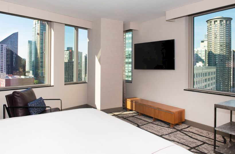 The image depicts a modern hotel room with large windows offering a cityscape view, a wall-mounted TV, a lounge chair, and minimalistic furniture.