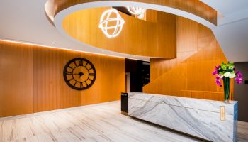 A modern interior features a marble reception desk, wooden staircase, large wall clock, and flower arrangement, under circular light fixtures.