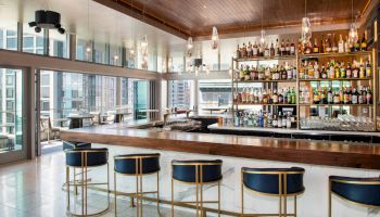 This image shows a modern, stylish bar with high stools, a wide selection of spirits, and large windows offering a city view.