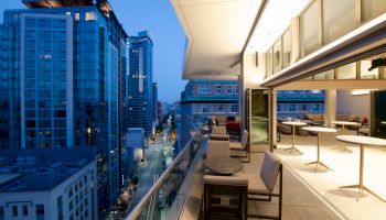 The image shows a modern cityscape from a building's balcony with tables, chairs, and a nighttime view of high-rise buildings and the street below.