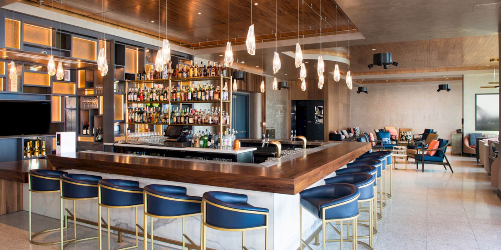 A modern bar with a wooden countertop, stylish barstools, pendant lights, and shelves filled with bottles, situated in a spacious, contemporary lounge.