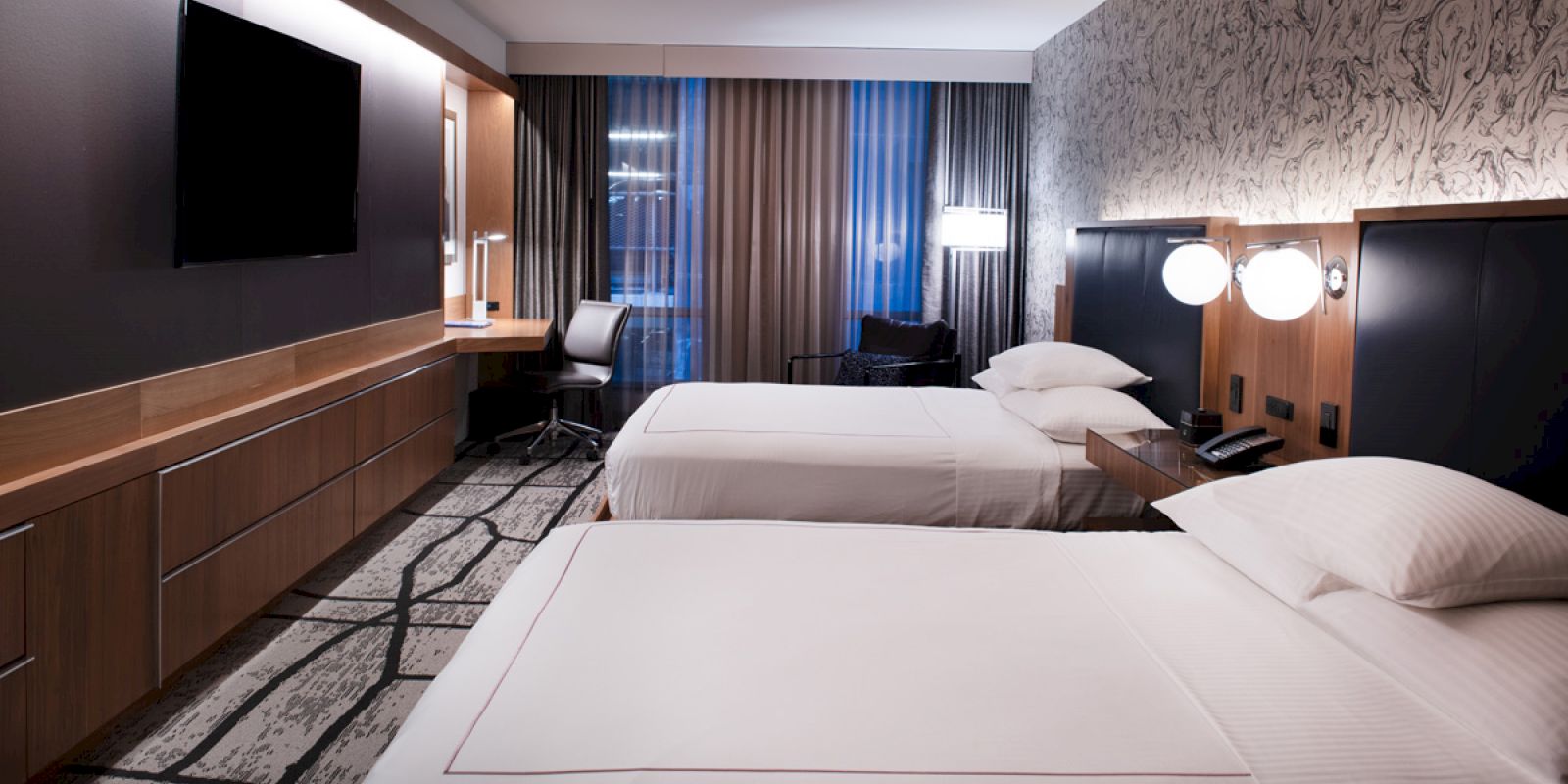 The image shows a modern hotel room with two beds, a flat-screen TV, a desk, and a large window with curtains. The room has ambient lighting.