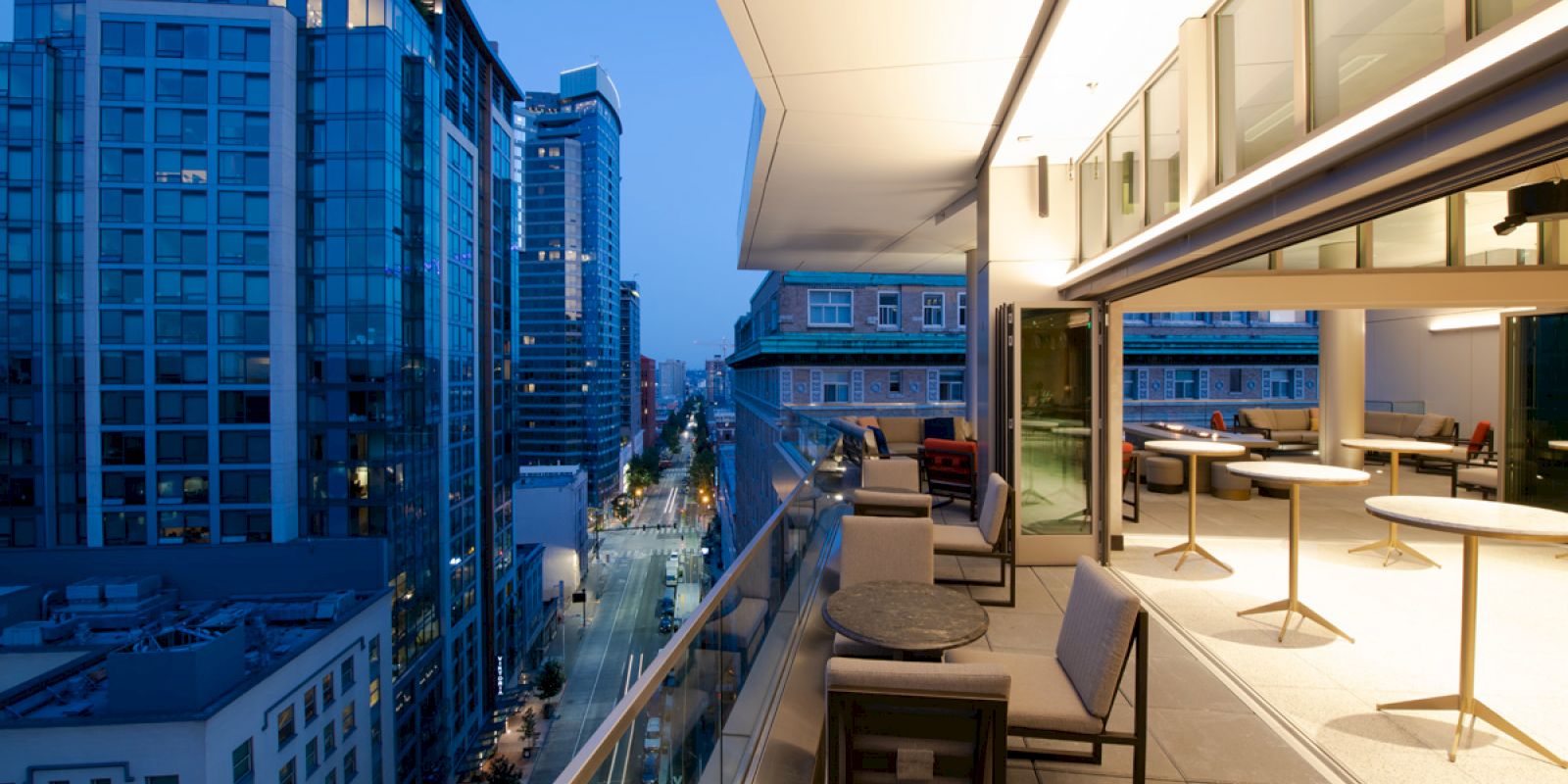 A modern cityscape view from a balcony with outdoor seating and tables, overlooking tall buildings and a street illuminated by evening lights.