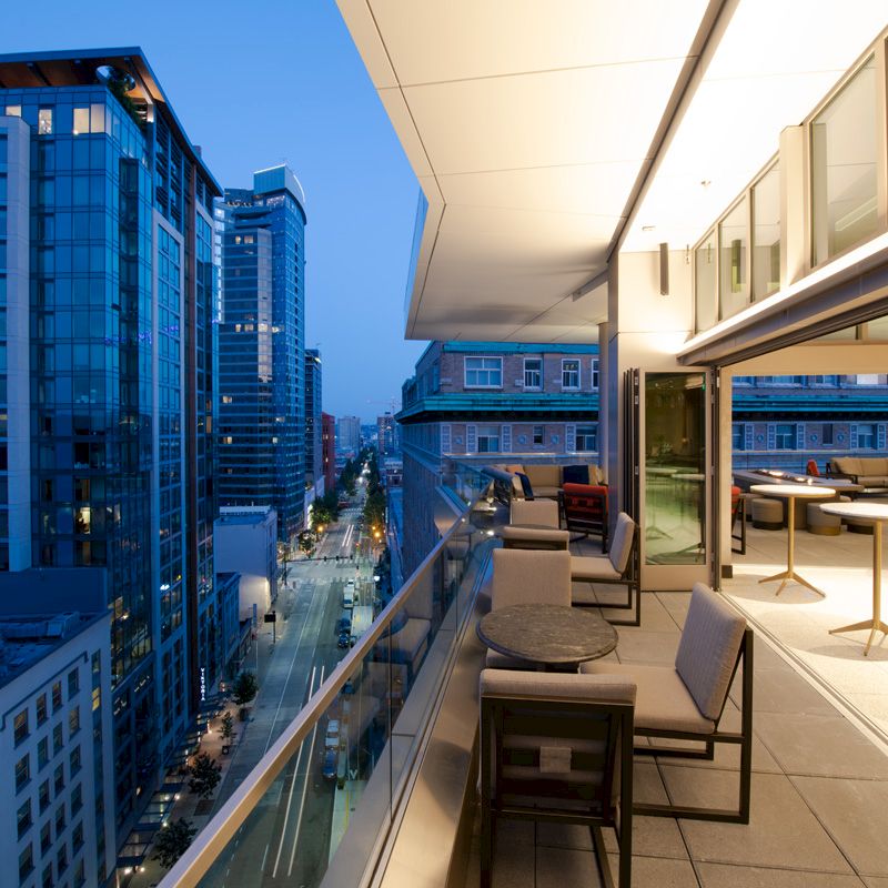 A modern balcony overlooks a cityscape at dusk. The balcony features outdoor seating, tables, and floor-to-ceiling windows with an indoor space.