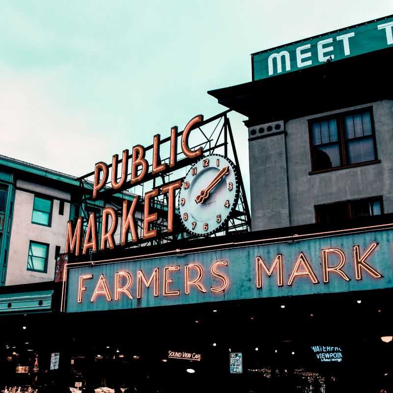 The image shows a neon sign for a Public Market and Farmers Market against a backdrop of buildings and a cloudy sky.