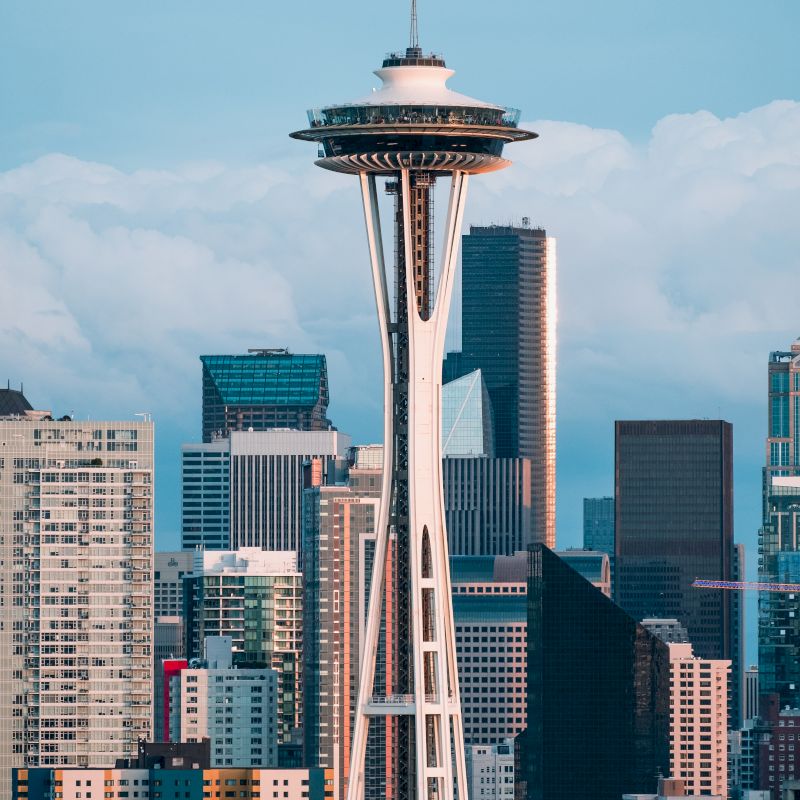 The image shows the iconic Space Needle with the Seattle skyline in the background, against a backdrop of clouds. The buildings are a mix of heights and styles.