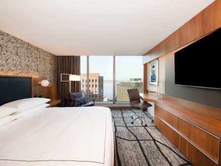 The image shows a modern hotel room with a bed, desk, large TV, chair by the window, and a view of city buildings through the window.