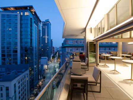An outdoor terrace with seating and tables overlooks a city street with tall buildings, captured during twilight with city lights visible.