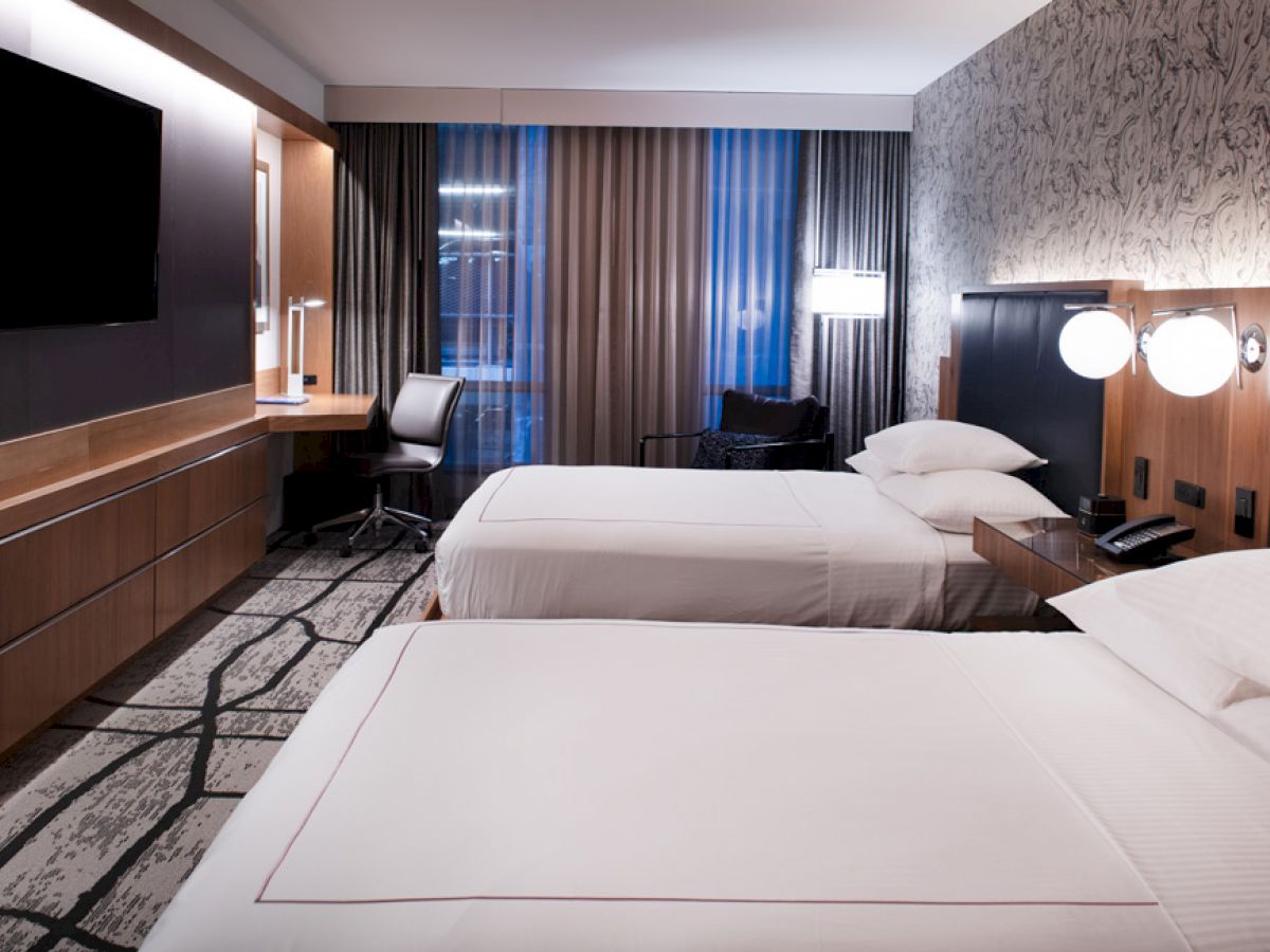 A hotel room with two double beds, a wall-mounted TV, a desk with a chair, and bedside lamps on a patterned wall.