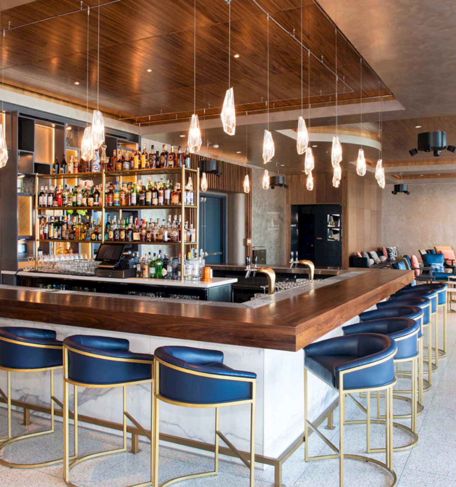 This image shows a modern bar with a well-stocked shelf of bottles, blue bar stools, hanging light fixtures, and additional seating in the background.