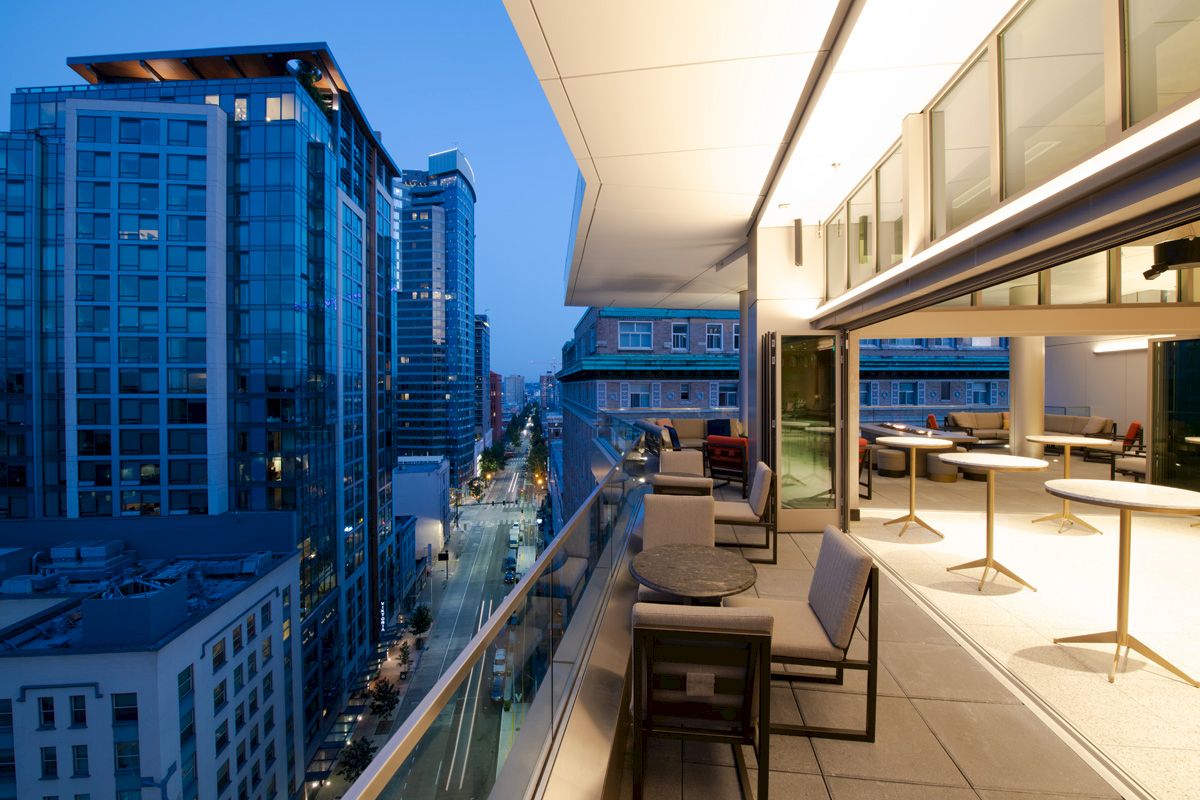 A modern cityscape at dusk, showing tall buildings and a well-lit, elegant balcony setup with sleek furniture and a glass railing.