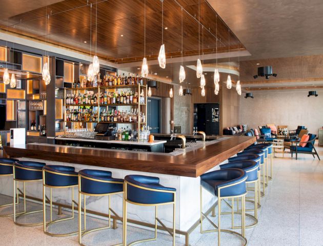 A modern bar with a well-stocked liquor shelf, wooden accents, stylish lighting, and blue bar stools. There are tables and seating in the background.